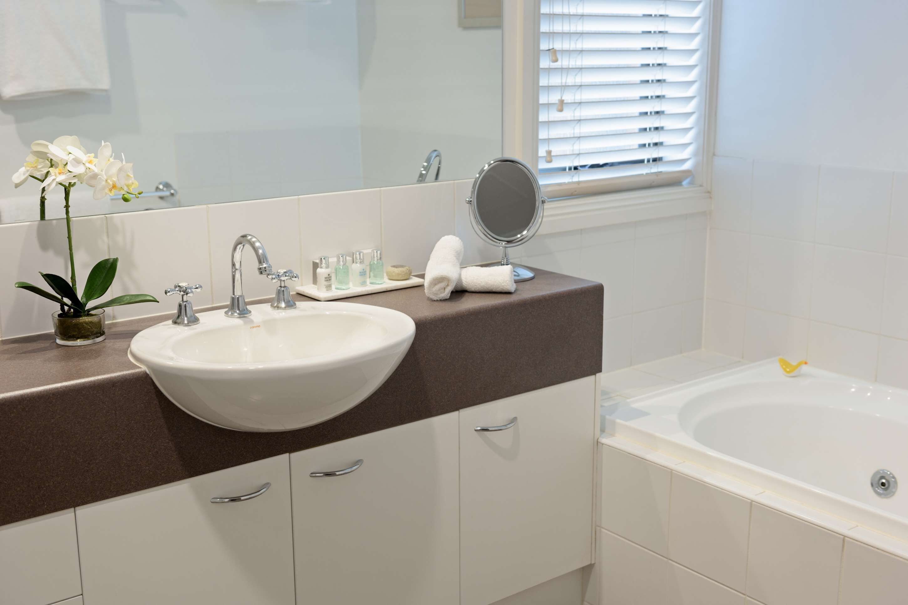 A bathroom with a white sink, a mirror, a bathtub, and a window with blinds.