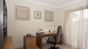 Una oficina en casa con escritorio, silla, impresora y archivador. Hay tres ventanas con persianas, una estantería y un ventanal con cortinas traslúcidas.