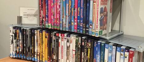A DVD rack with various movie titles and a window with blinds in the background.