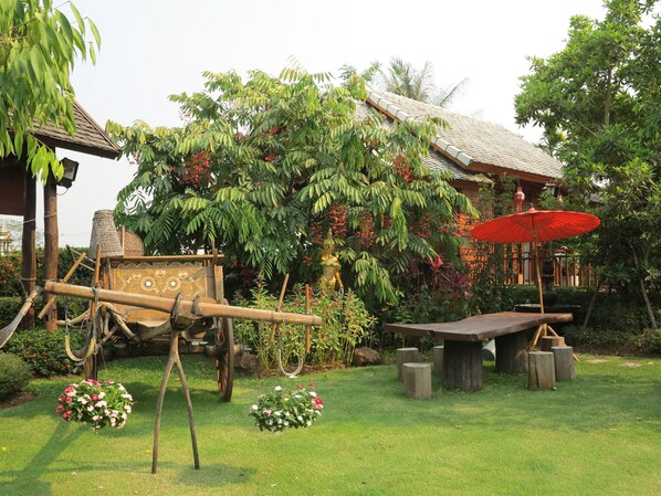 Un carruaje de madera tradicional con detalles decorativos, un paraguas rojo y una mesa de piedra en un jardín exuberante lleno de plantas, con un edificio de techo de tejas al fondo.