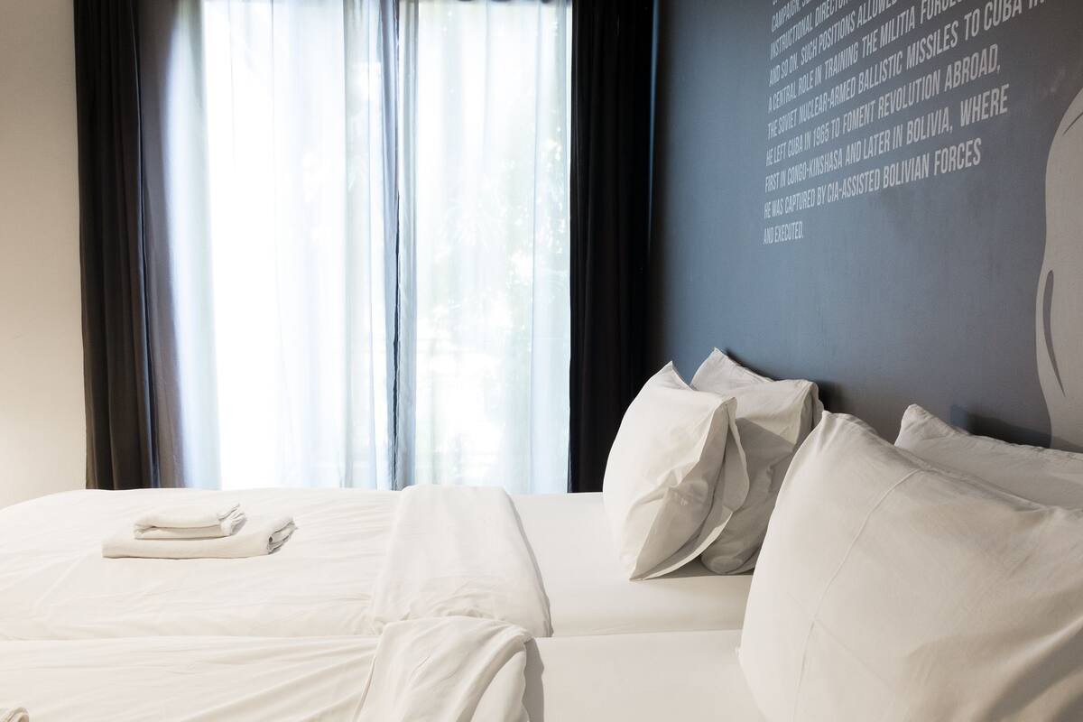 Habitación de hotel con una cama grande, ropa de cama blanca y vistas al exterior a través de cortinas traslúcidas.