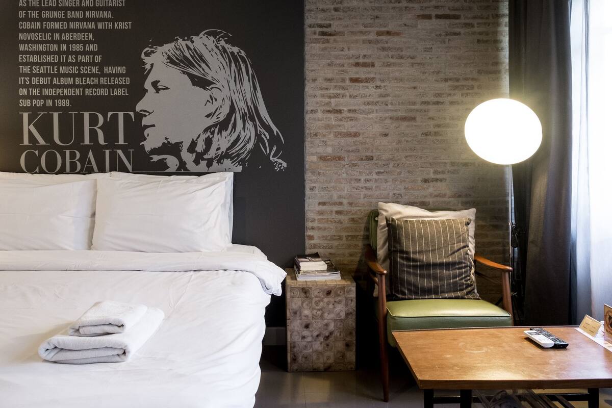 Habitación de hotel con cama, mesita de noche, una silla, una mesa y una pared con un mural de un músico.