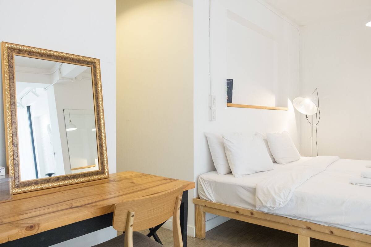 Un dormitorio con un escritorio de madera, una cama con ropa blanca, un espejo grande con marco dorado y una lámpara de pie.
