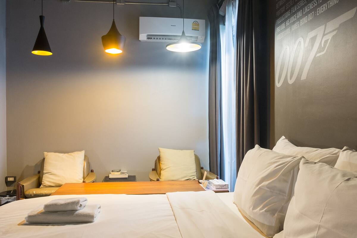 Habitación de hotel con una cama grande, dos mesitas de noche, un escritorio y una silla. Se observan dos lámparas colgantes, un equipo de aire acondicionado y una ventana con cortinas.