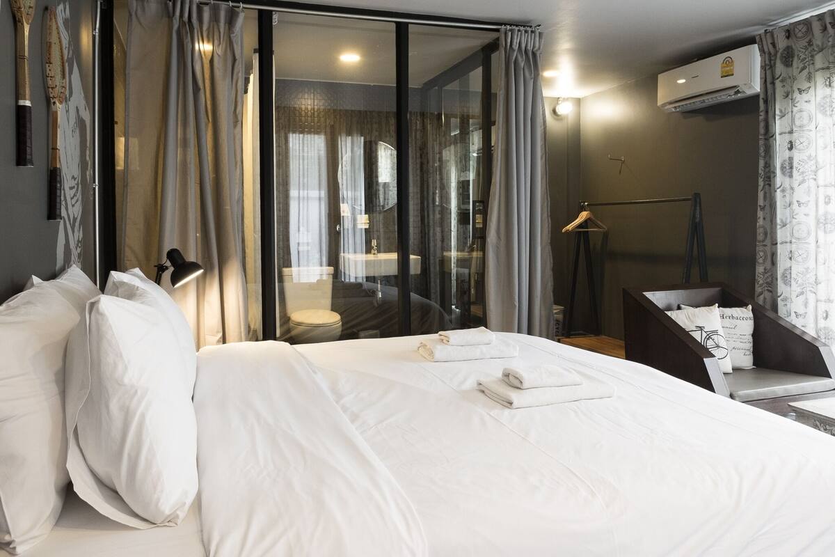Una habitación de hotel moderna con una cama grande, un separador de vidrio, un baño y vistas de la ciudad.