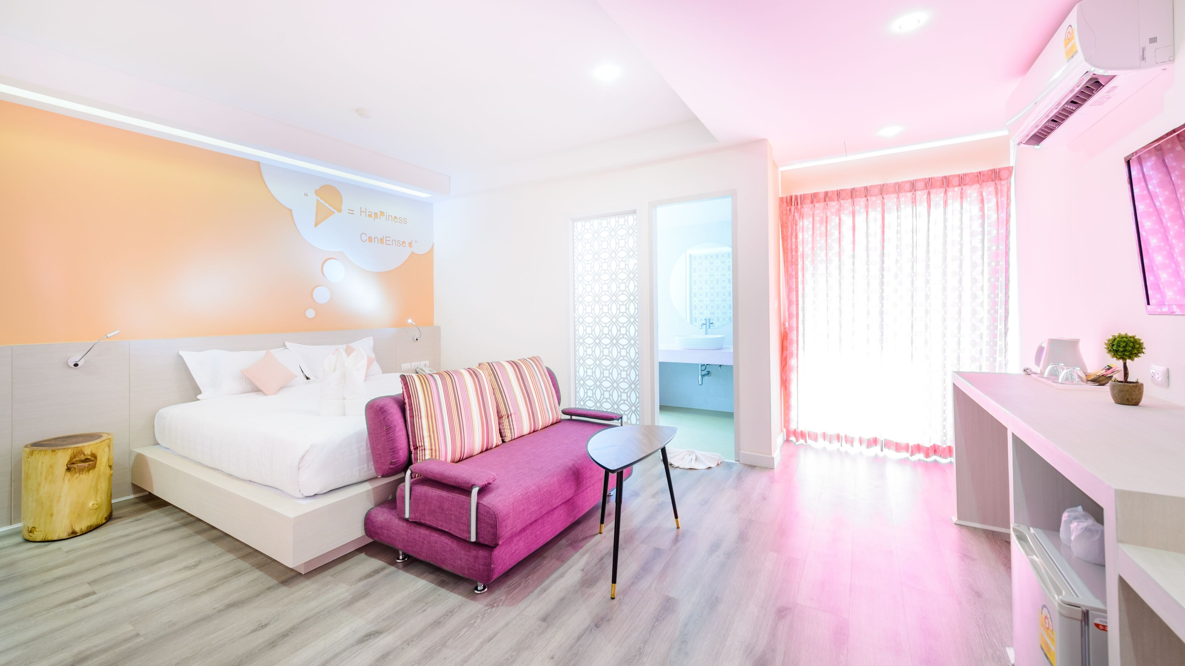 Un dormitorio moderno con cama, un sofá púrpura, una mesita pequeña y un baño visible a través de una puerta de cristal.