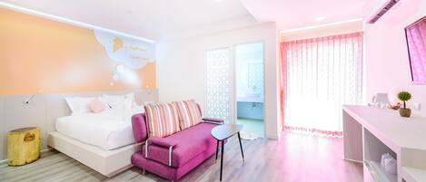 Un dormitorio moderno con cama, un sofá púrpura, una mesita pequeña y un baño visible a través de una puerta de cristal.