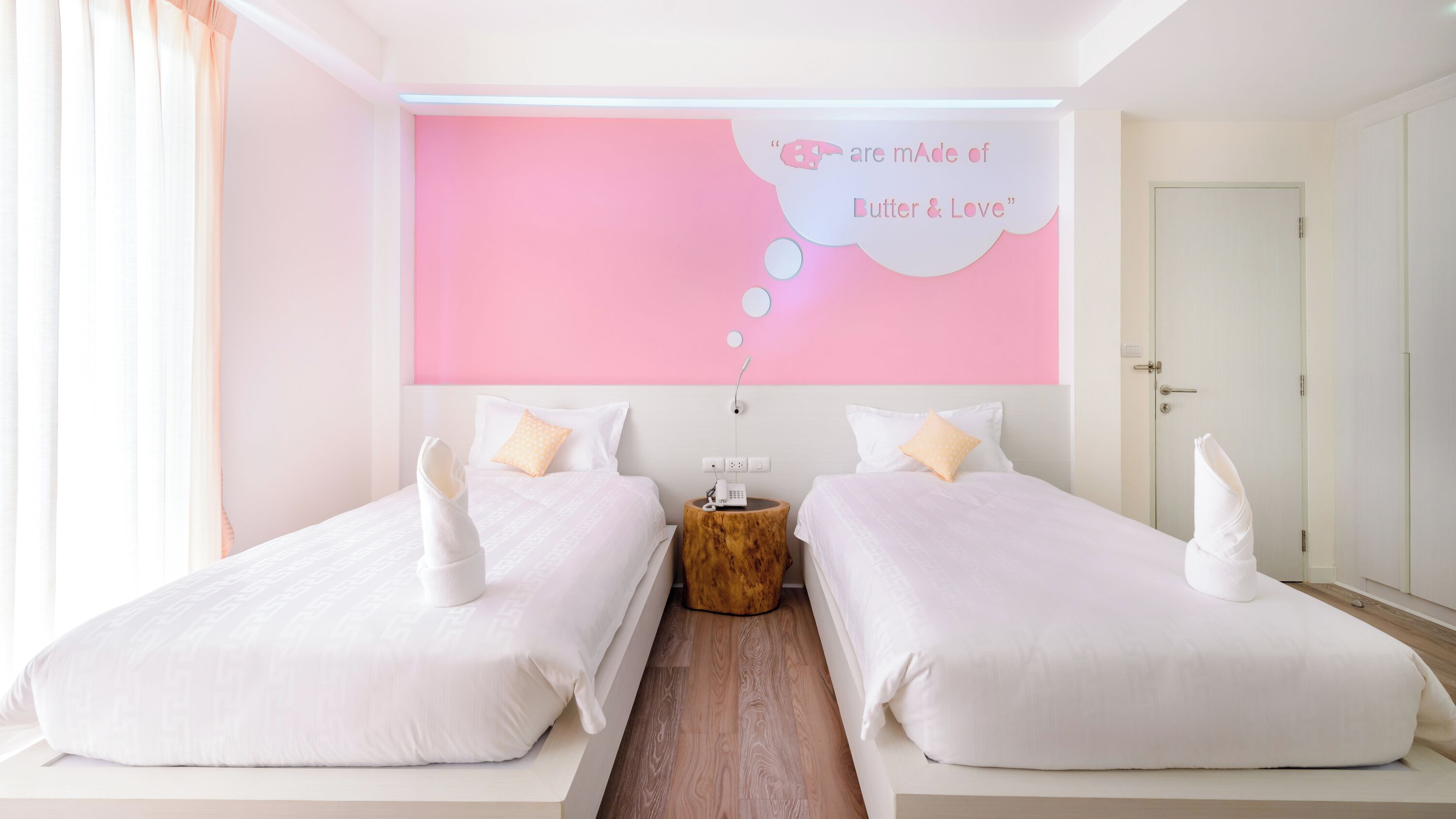 A hotel room with two beds, a wooden nightstand, a wall-mounted phone, and a pink accent wall with a quote.