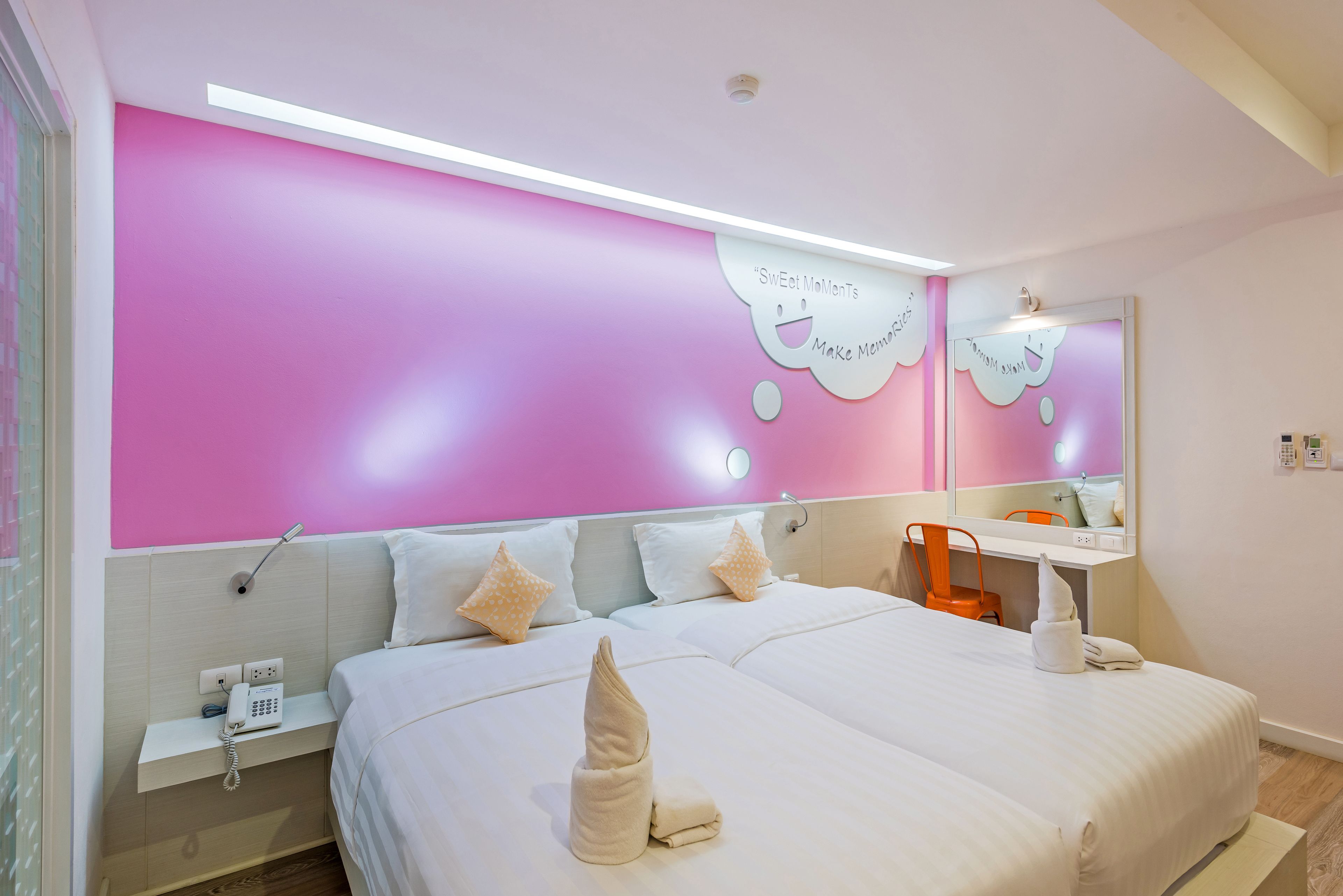 A hotel room with a pink accent wall, a bed with white bedding, a bedside table with a phone, and a mirror above the bed.