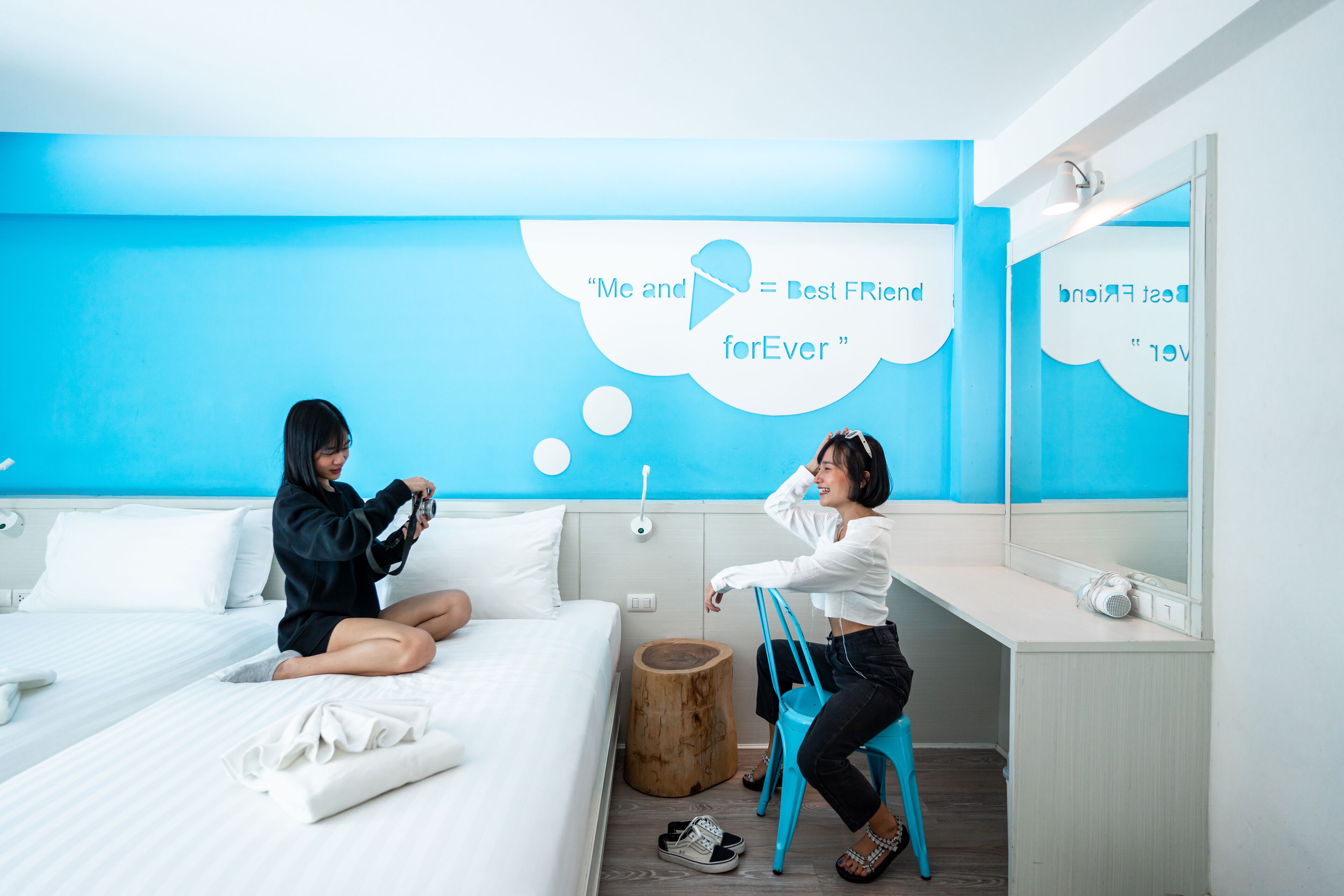 A modern hotel room with a blue accent wall, a bed with white linens, a wooden stool, and a mirror reflecting the room.