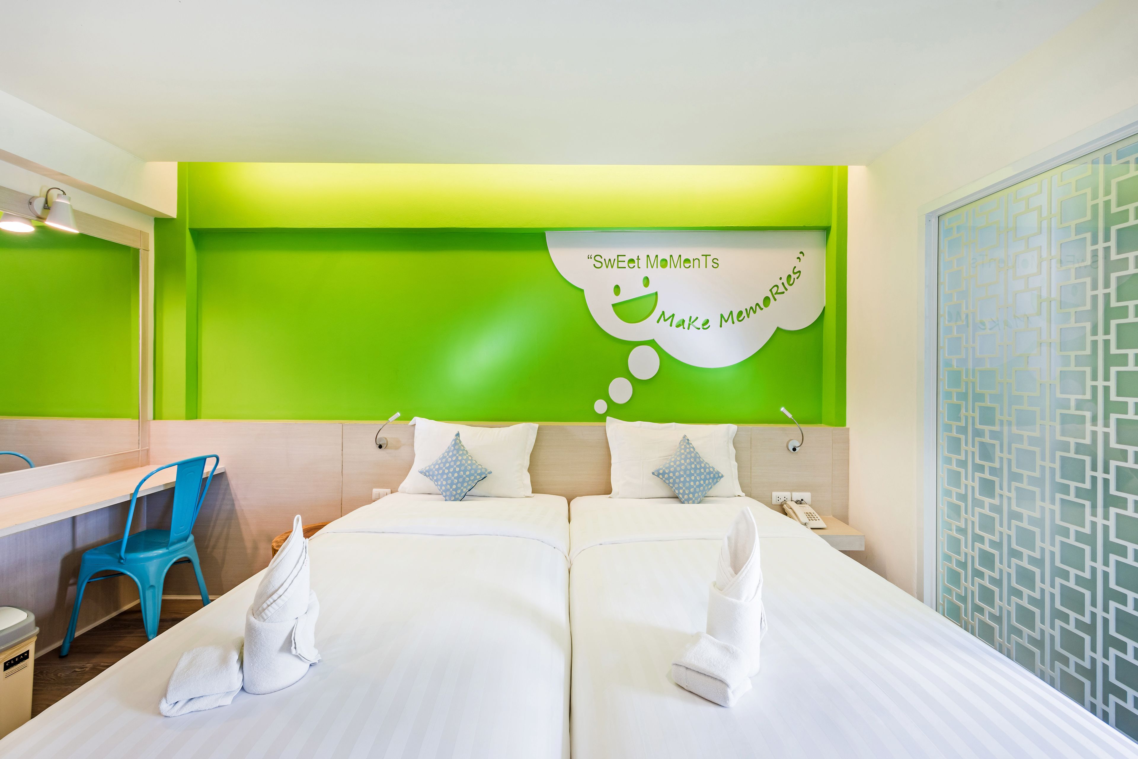 Et hotelværelse med to senge, en blå stol, et spejl og en væg med grønne detaljer samt en taleboble med teksten "Sweet MoMenTs Make Memories".