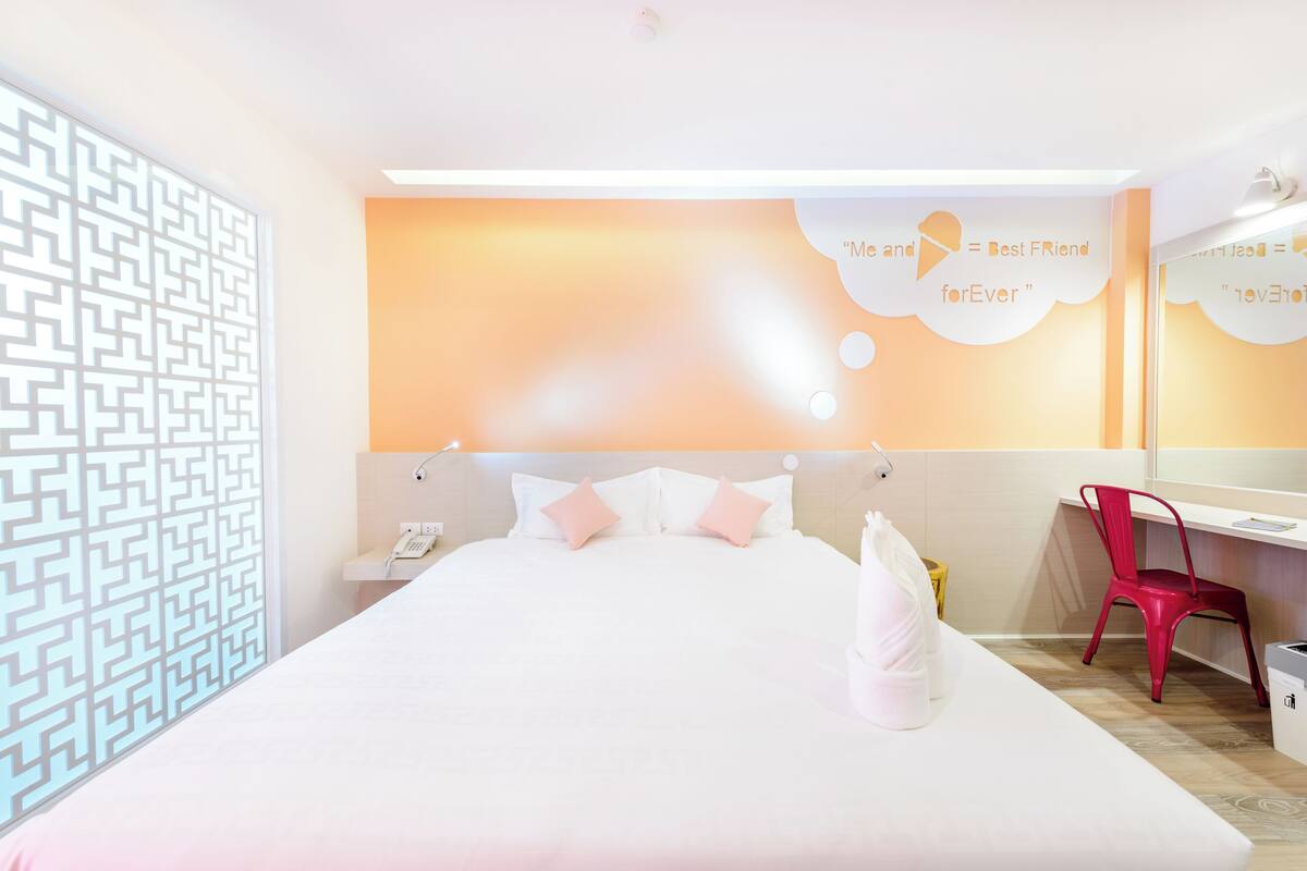 Una habitación de hotel moderna con una cama grande, un escritorio y una silla. Hay una pared decorativa con un diseño geométrico y una cita sobre helado.