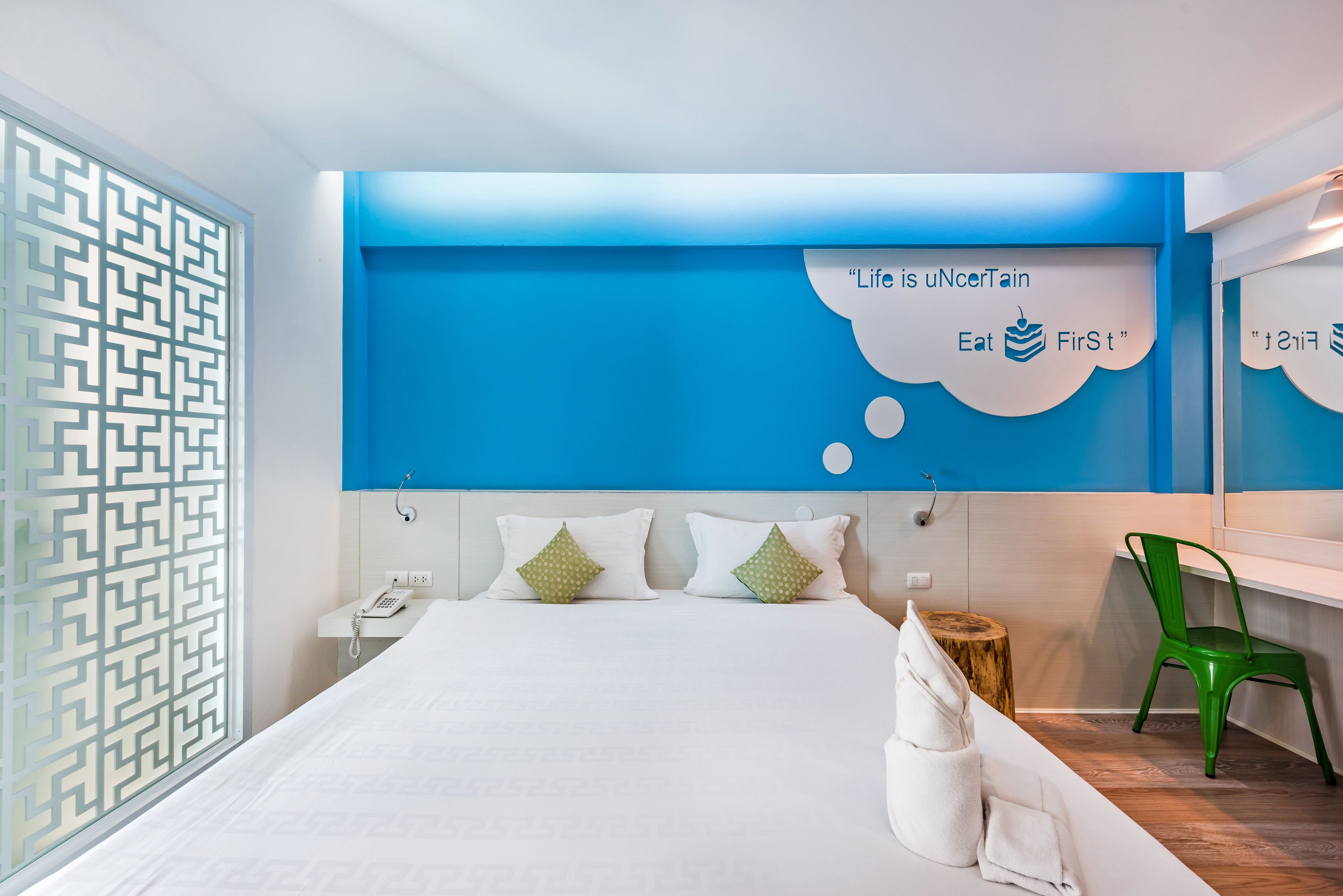Una habitación de hotel moderna con una cama grande, un teléfono en la mesita de noche, una silla verde y una pared con detalles azules y una burbuja de pensamiento que dice 'Life is unCertain Eat First'.