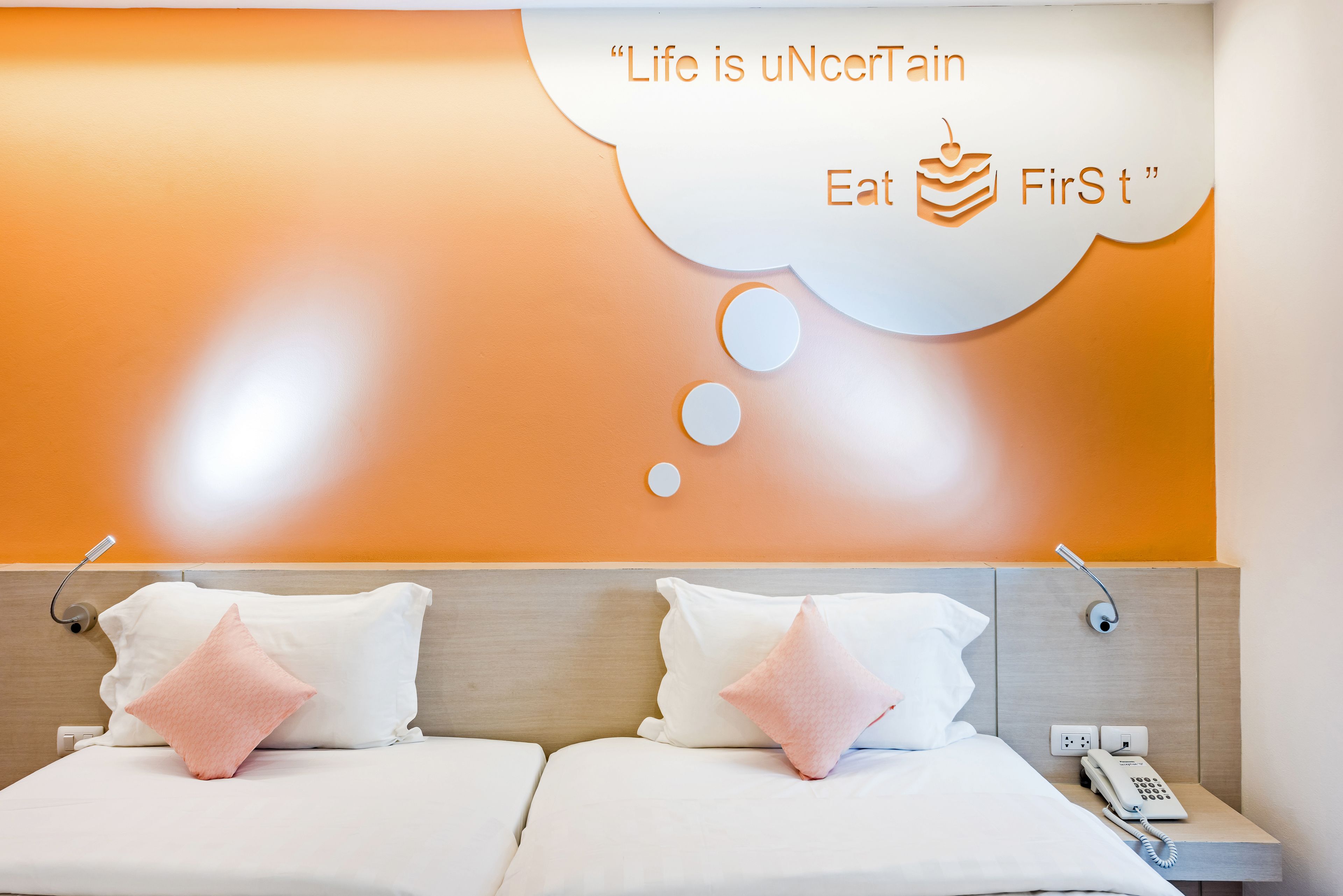 Habitación de hotel con dos camas, cada una con ropa blanca y almohadas rosas. Hay un teléfono montado en la pared y una cita que dice "Life is uNcErTain Eat FirSt".