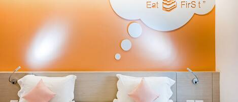 Habitación de hotel con dos camas, cada una con ropa blanca y almohadas rosas. Hay un teléfono montado en la pared y una cita que dice "Life is uNcErTain Eat FirSt".