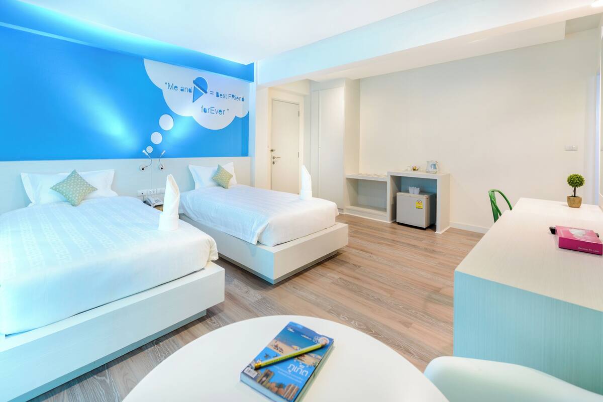 Una habitación de hotel moderna con dos camas, un escritorio, una silla, una mesita y una pared con detalles azules y un gráfico de bocadillo de diálogo.