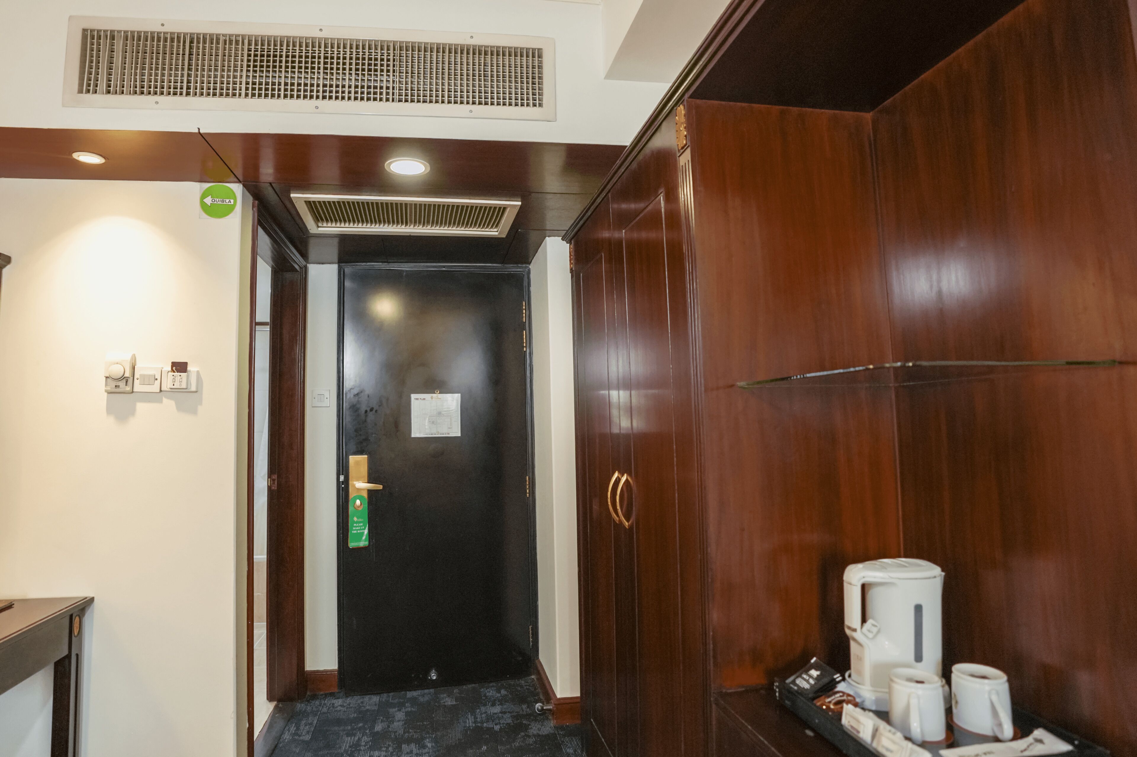 A hotel entrance with a dark wooden door, a green directional sign, a coffee station with cups and a kettle, and a vent on the ceiling.