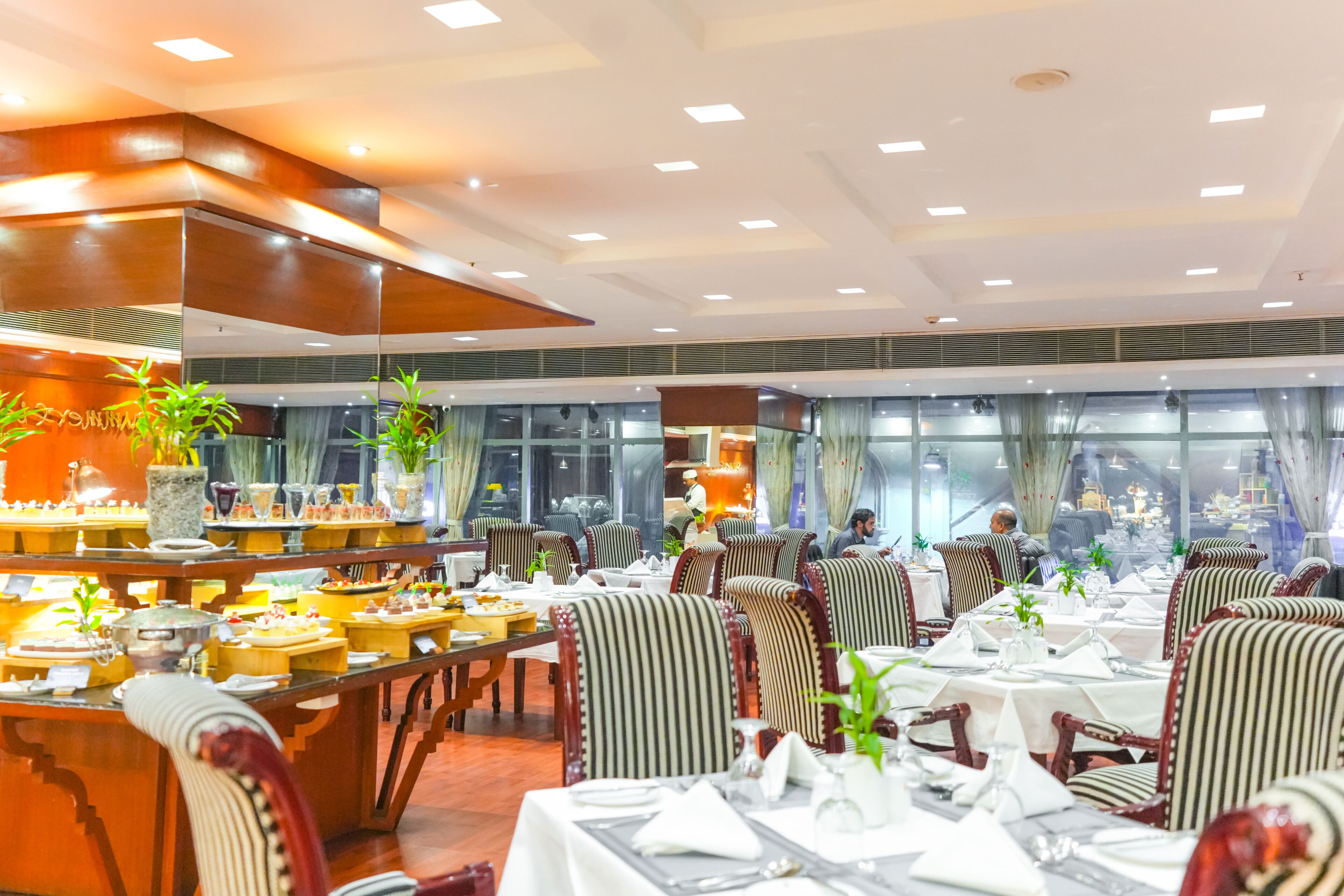 Ein Restaurant mit mehreren Tischen und Stühlen, einem Küchenbereich im Hintergrund und großen Fenstern mit Vorhängen.