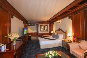 Habitación de hotel con una cama grande, televisor, escritorio con un florero, una silla y una mesita de noche con lámpara.