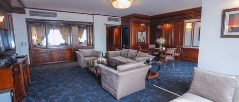 A spacious living room with a large sofa, a coffee table, and a fireplace. There is a large window with curtains, a chandelier, and a wooden cabinet.