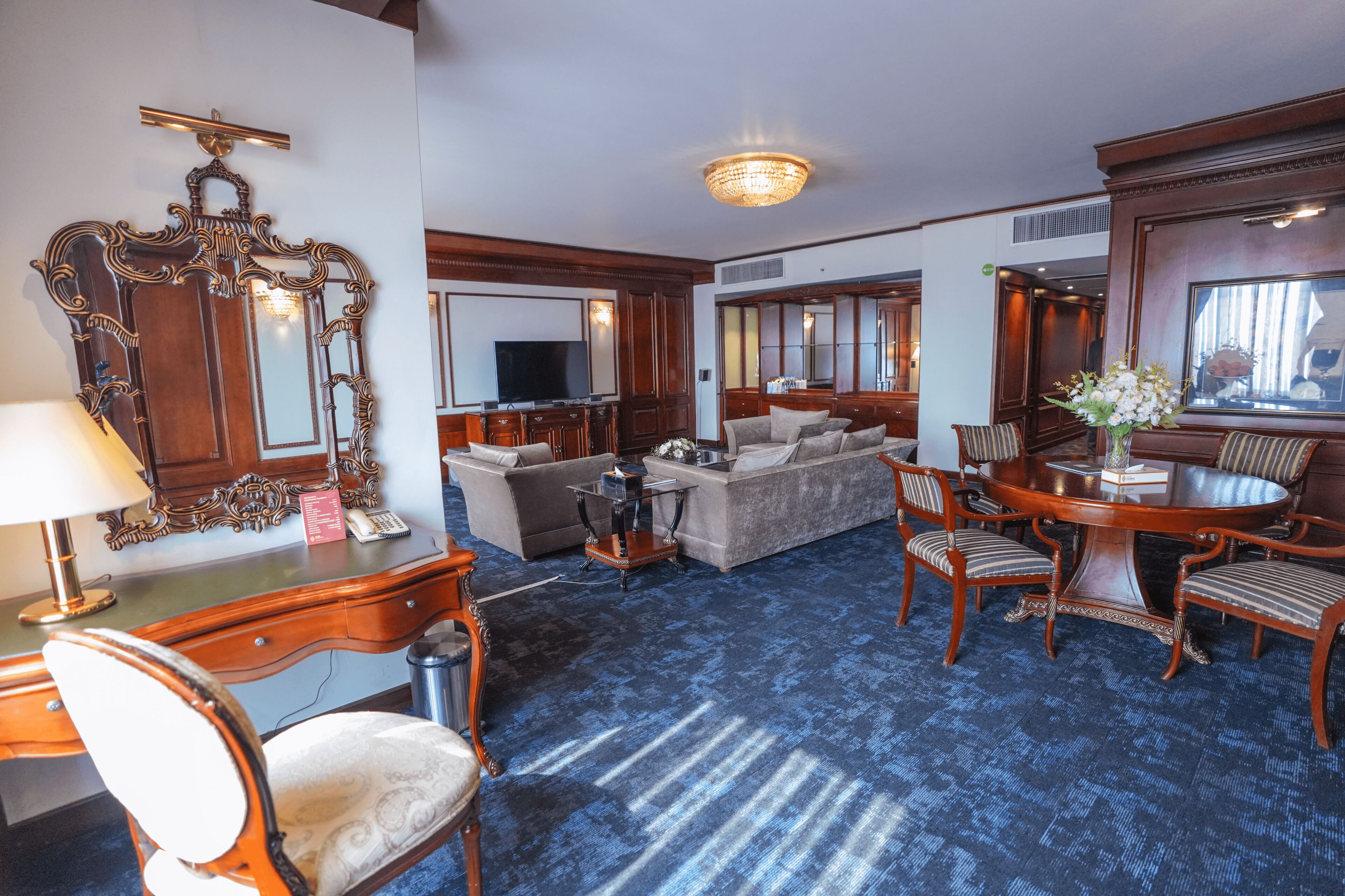 A hotel room with a blue carpet, a wooden desk with a lamp, a sofa, a dining table with chairs, and a large mirror.