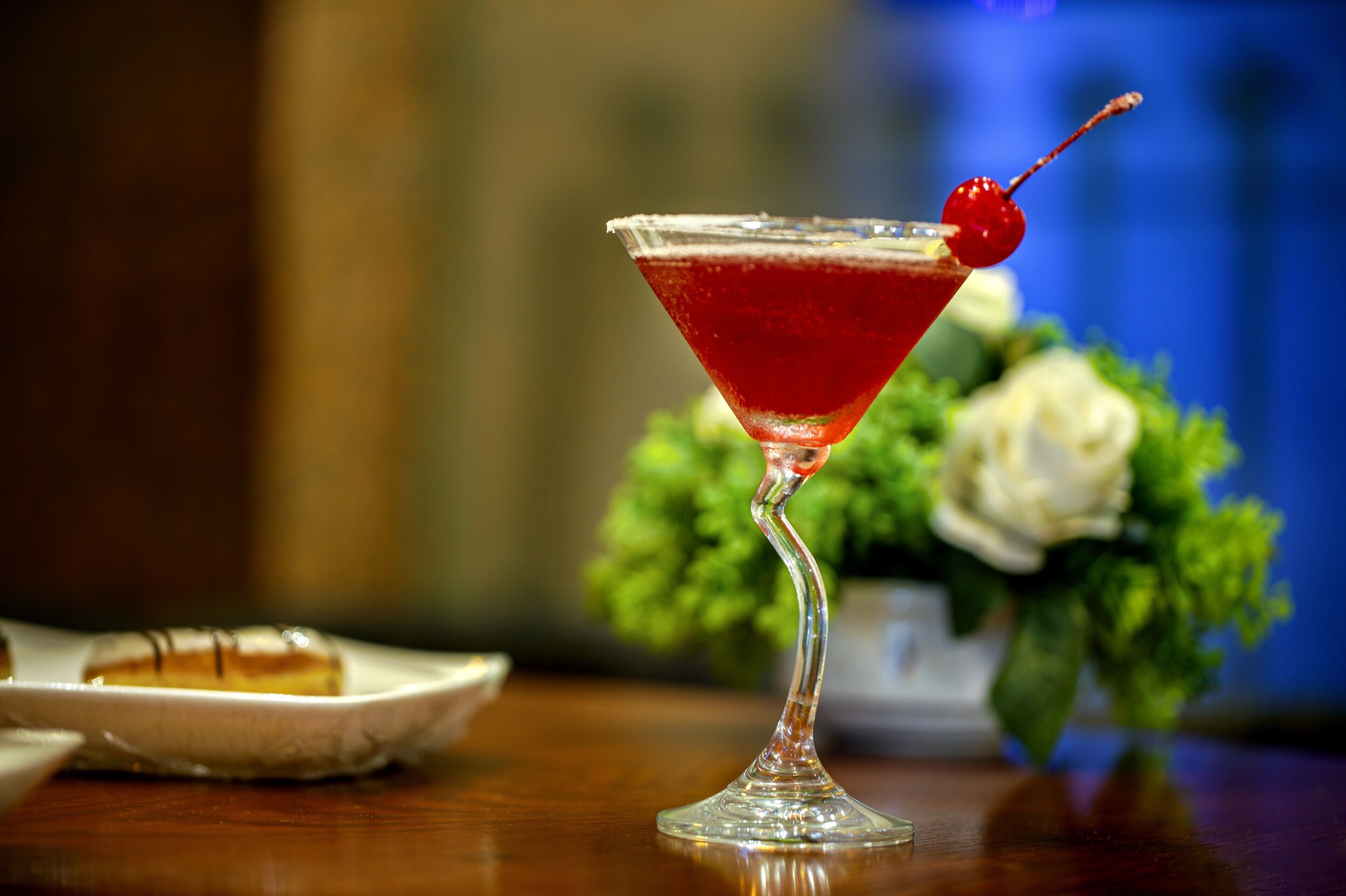 Un bicchiere da martini con una ciliegina sullo stelo, appoggiato su un tavolo di legno con sullo sfondo sfocato vegetazione e un'elegante composizione floreale bianca.