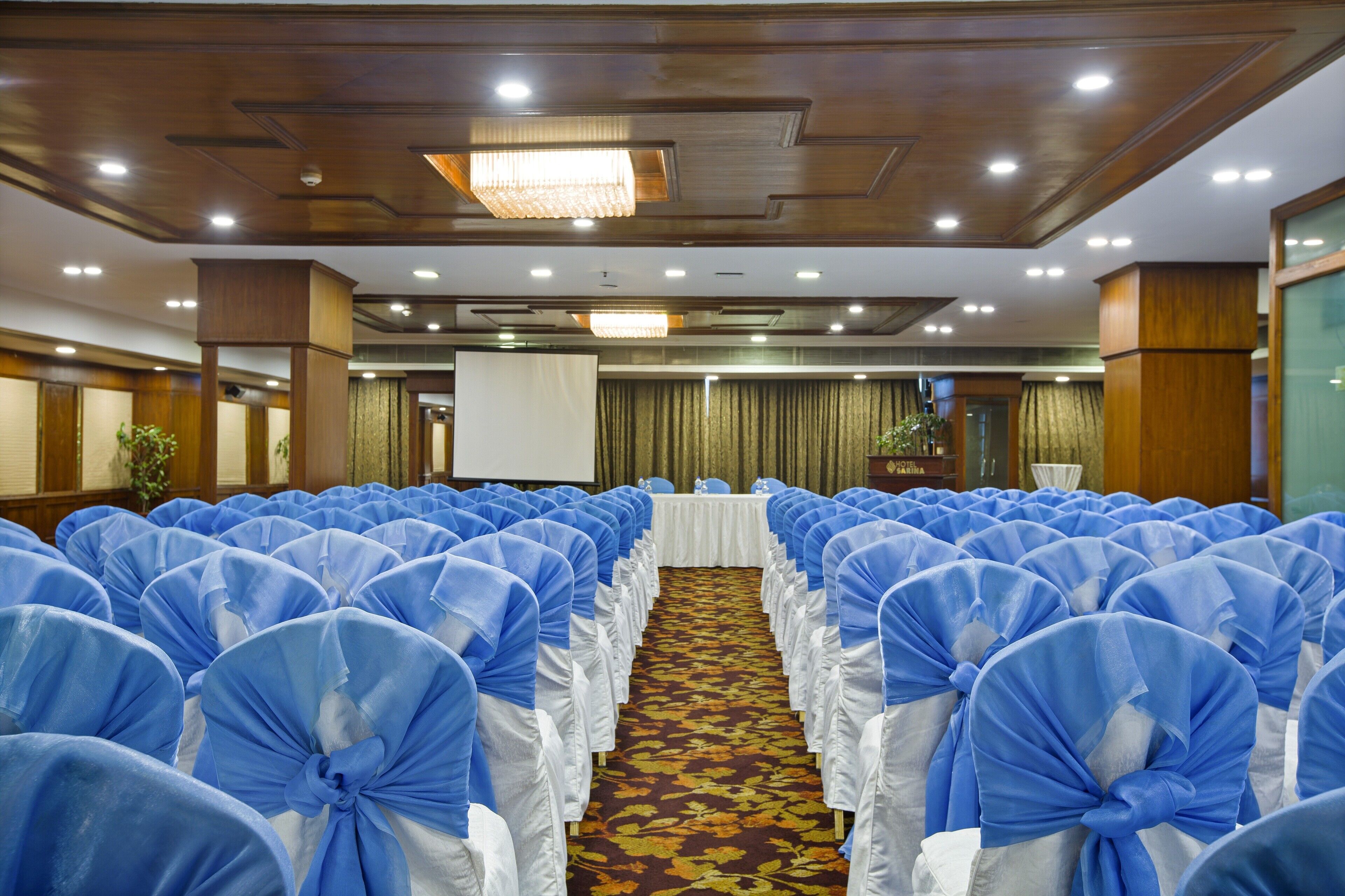 A conference room with rows of chairs covered in blue and white fabric, a white table at the front, and a carpeted floor.