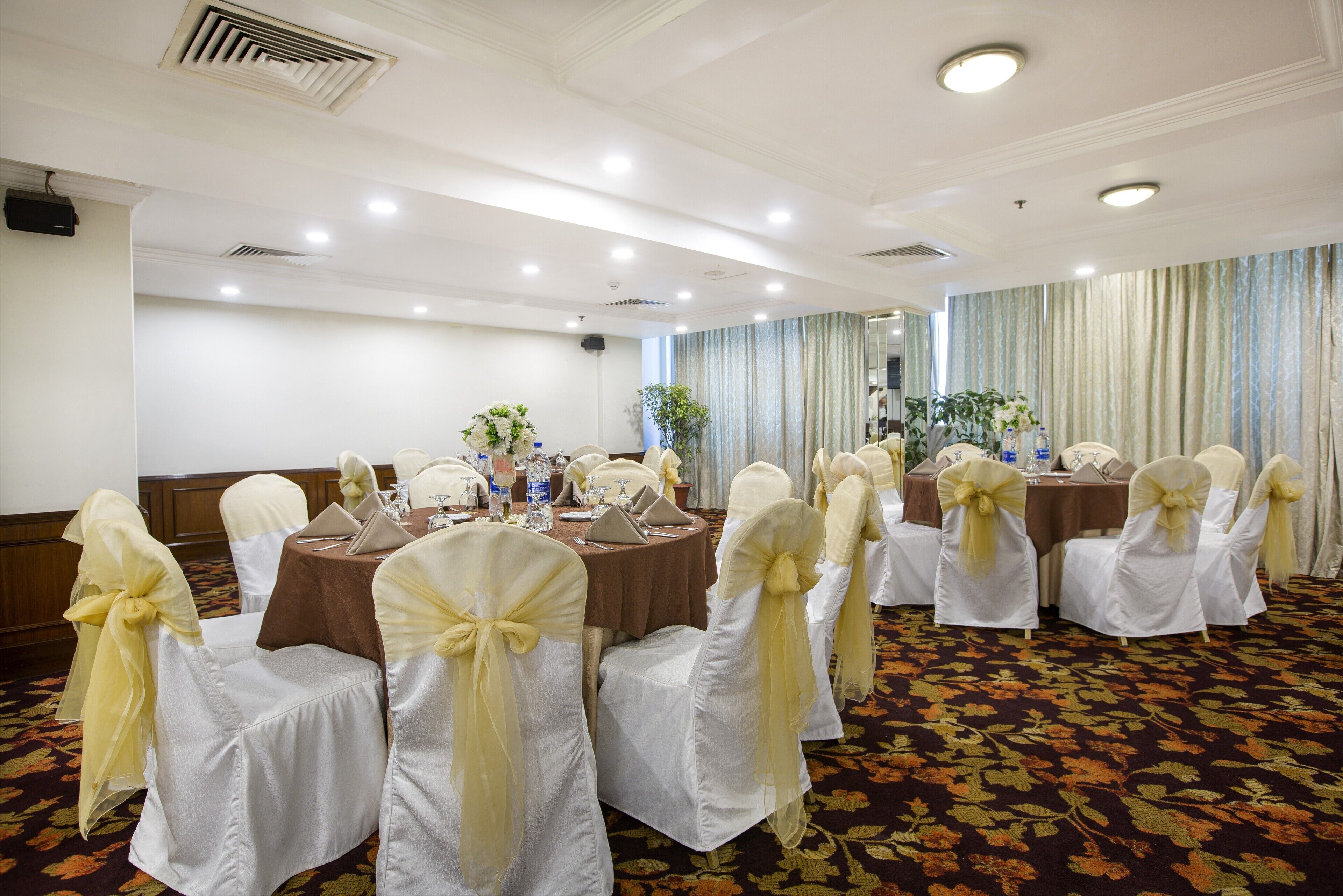 Un salón de banquetes con mesas redondas, sillas blancas con cintas amarillas y centros de mesa florales.
