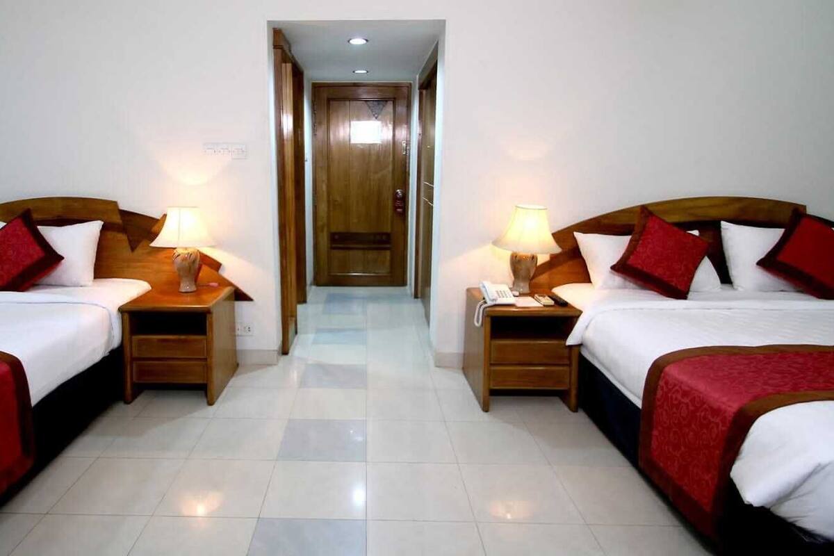 A hotel room with two beds, each with a wooden headboard, white bedding, and red accents. There are bedside tables with lamps and a visible door in the background.