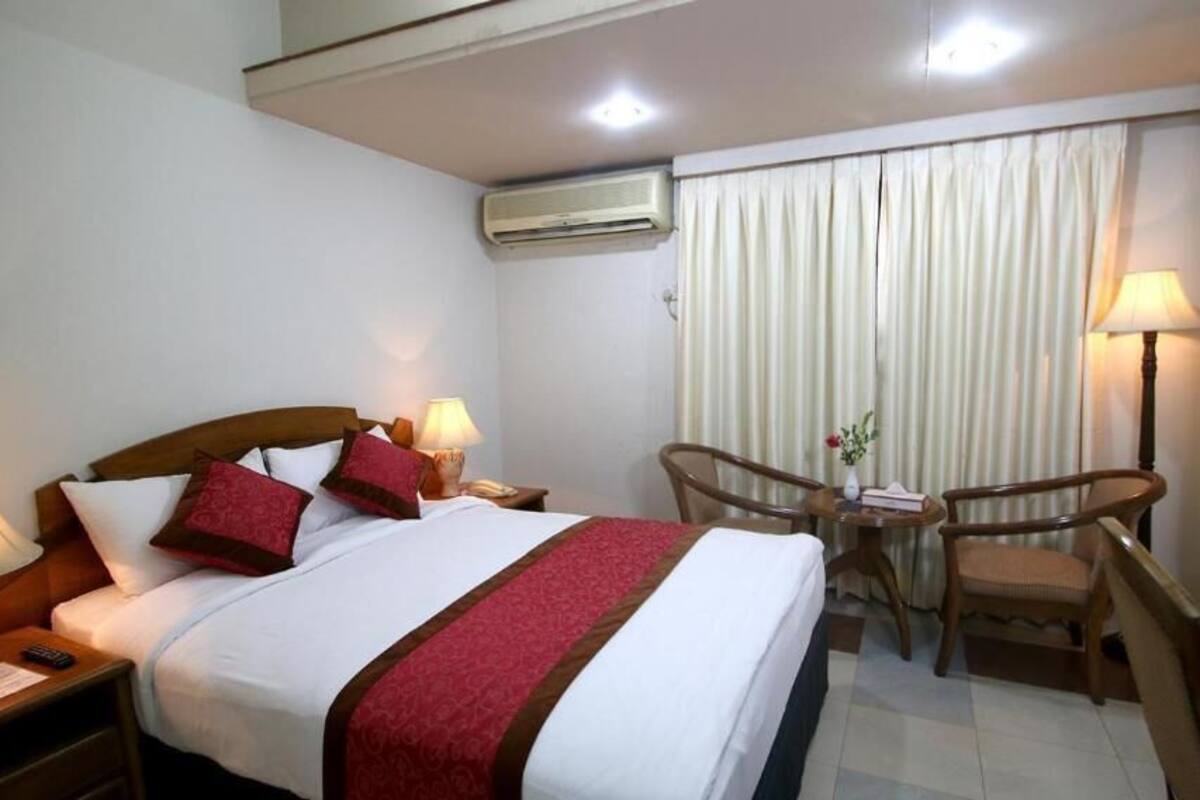 A hotel room with a bed, two bedside tables with lamps, a chair, a small table, and a window with curtains.