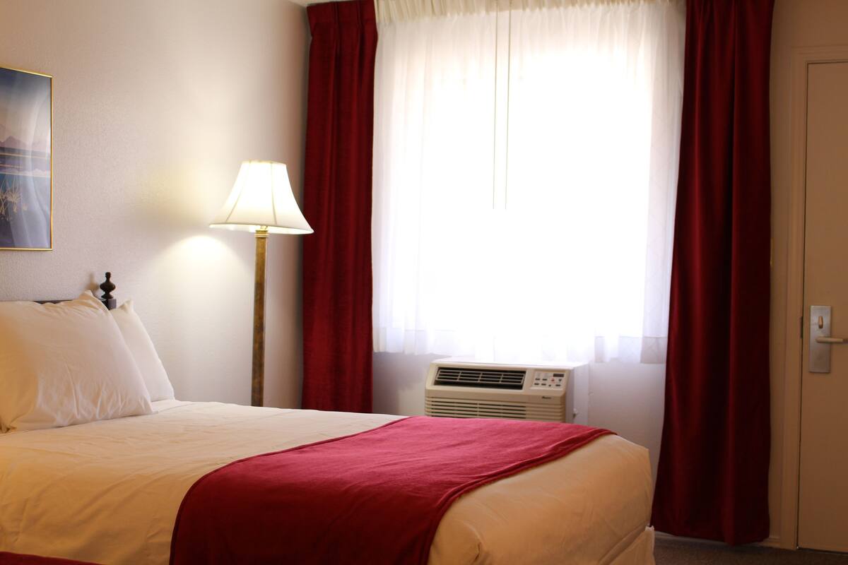 A hotel room with a bed, a lamp, an air conditioner, and a window with curtains.