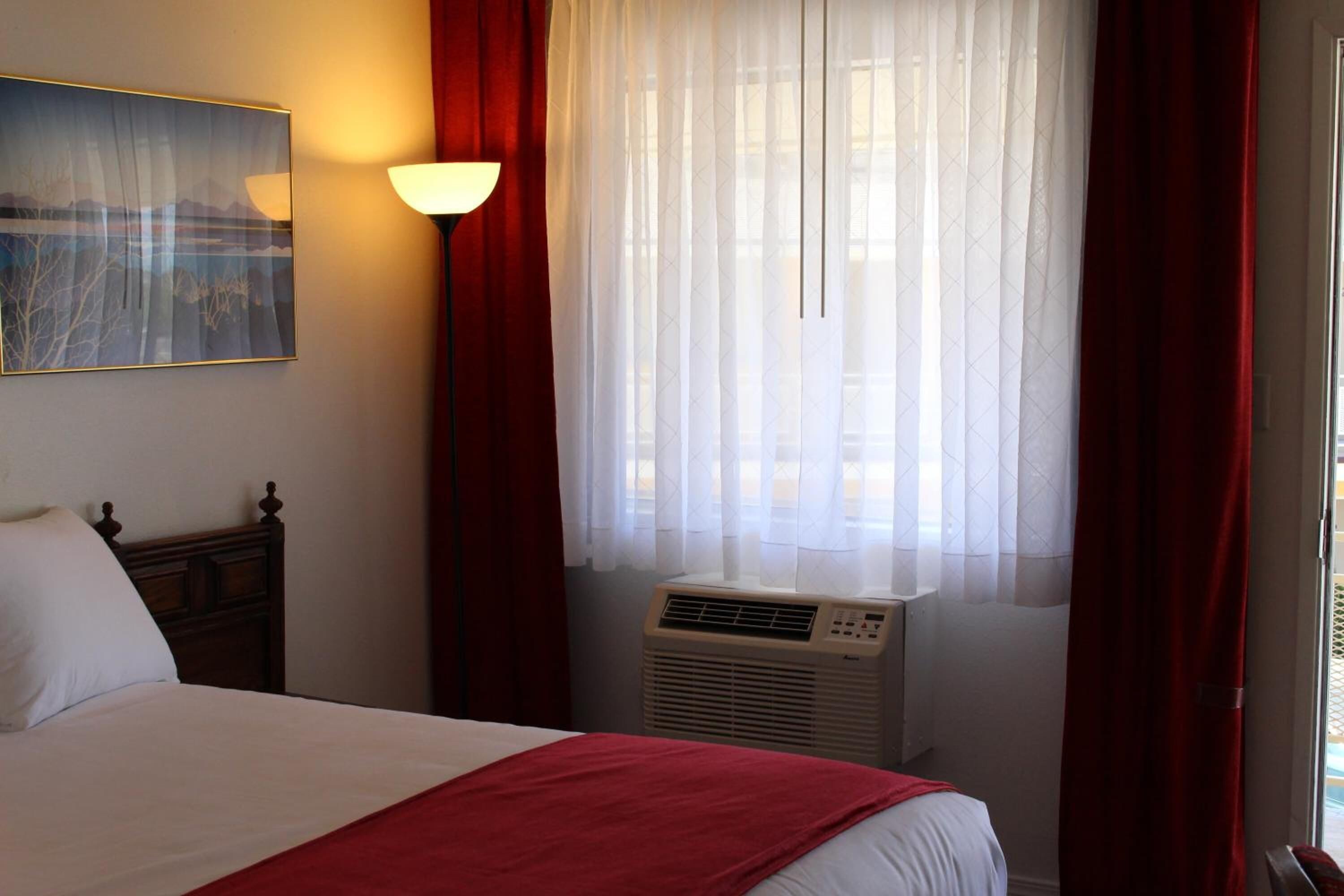 Habitación de hotel con cama, aire acondicionado de pared, ventana con cortinas traslúcidas y un cuadro en la pared.
