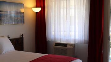 Habitación de hotel con cama, aire acondicionado de pared, ventana con cortinas traslúcidas y un cuadro en la pared.