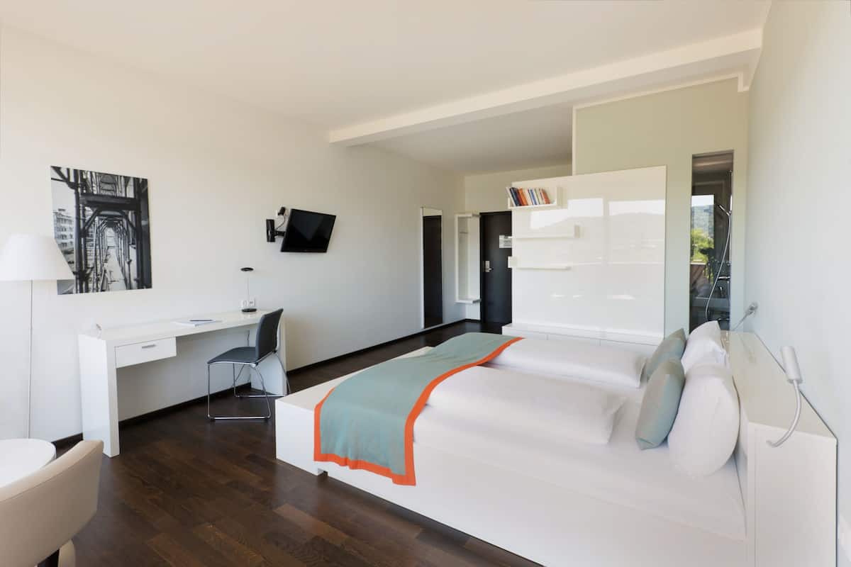 A modern hotel room with a bed, a desk, a chair, a television, and a bookshelf.
