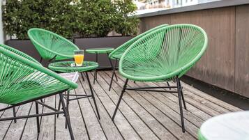 Eine Holzterrasse mit grünen Korbstühlen und einem kleinen Tisch, ein Glas Orangensaft und eine Holzwand im Hintergrund.