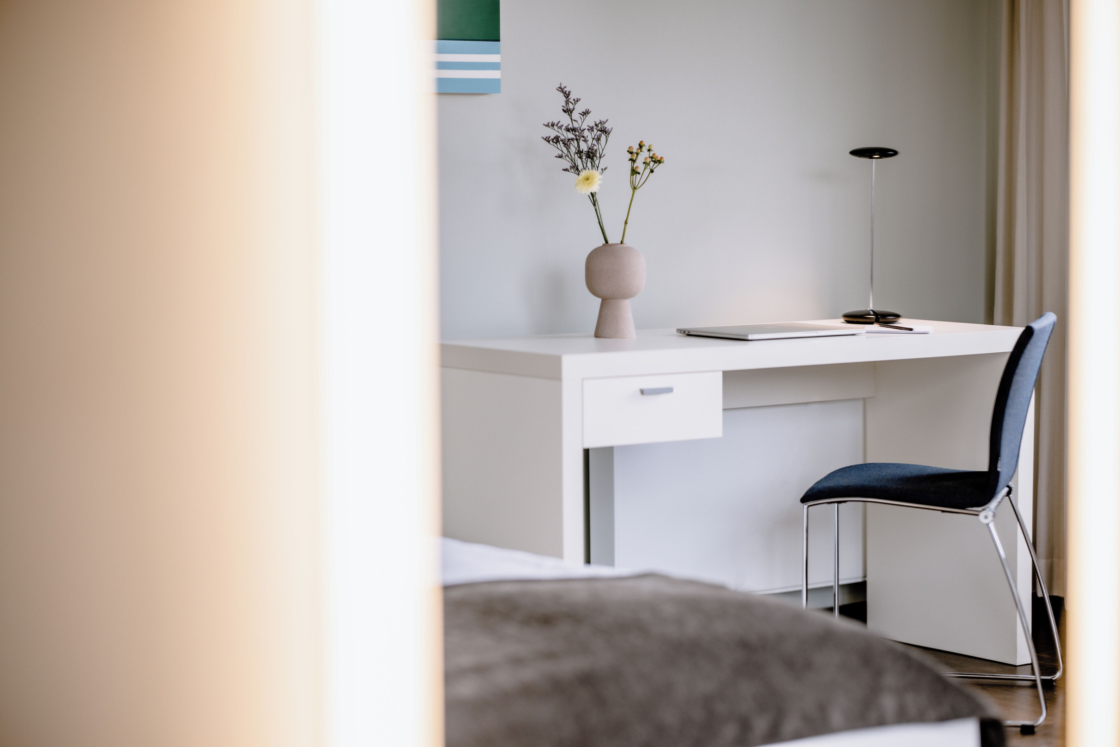 A modern hotel room with a white desk, a blue chair, a vase with flowers, and a floor lamp.