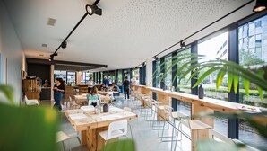 Un café moderno con mesas y sillas de madera, ventanas amplias y plantas de interior.