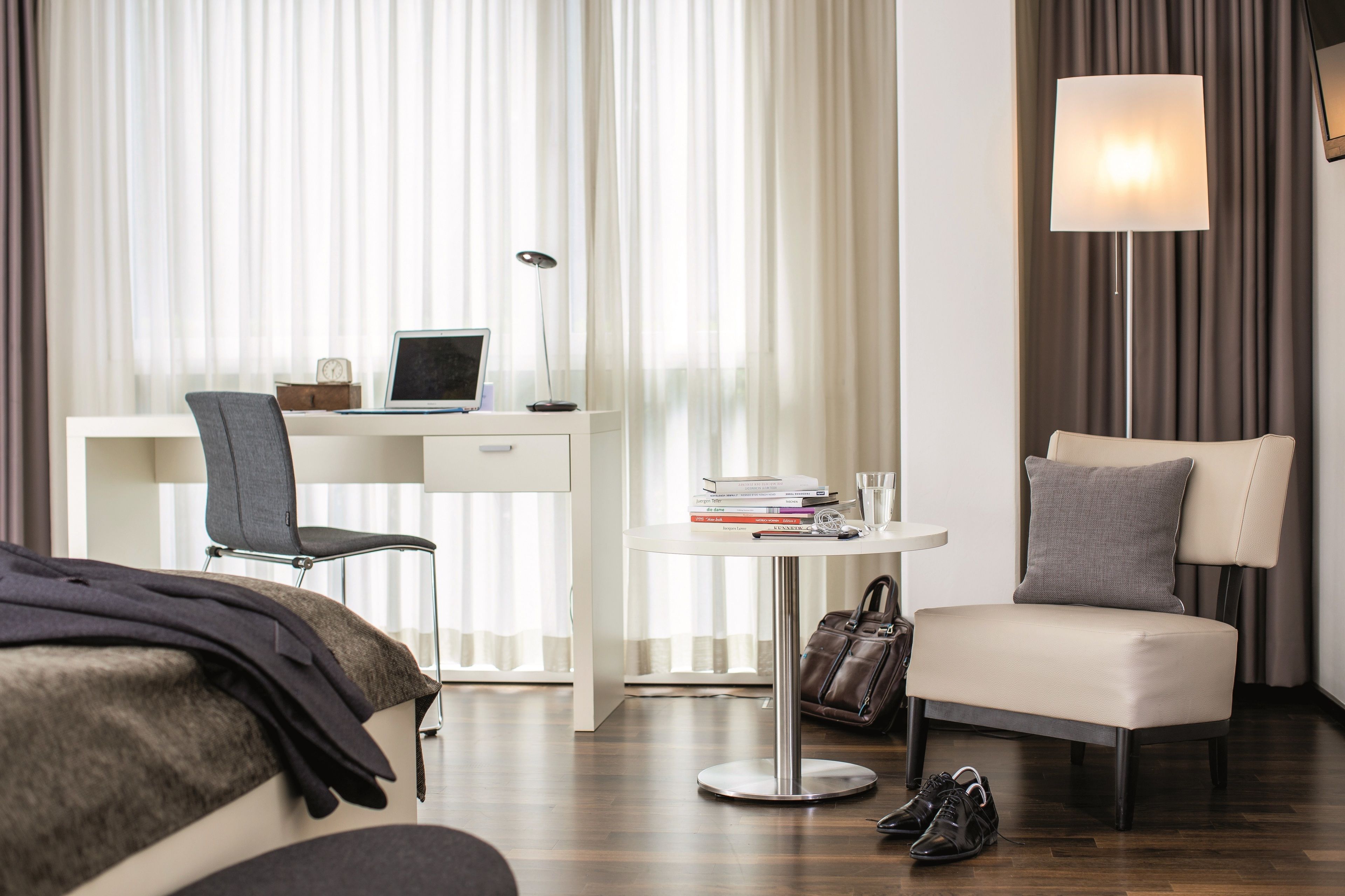 A hotel room with a bed, a desk with a laptop, a chair, a small table with books and a lamp, and a bedside table with a handbag and shoes.