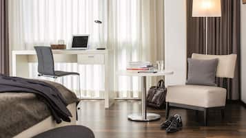 A hotel room with a bed, a desk with a laptop, a chair, a small table with books and a lamp, and a bedside table with a handbag and shoes.