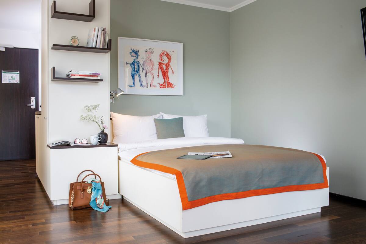 A neatly arranged hotel room with a bed, bedside table, and a wall-mounted shelf with books and a framed picture.