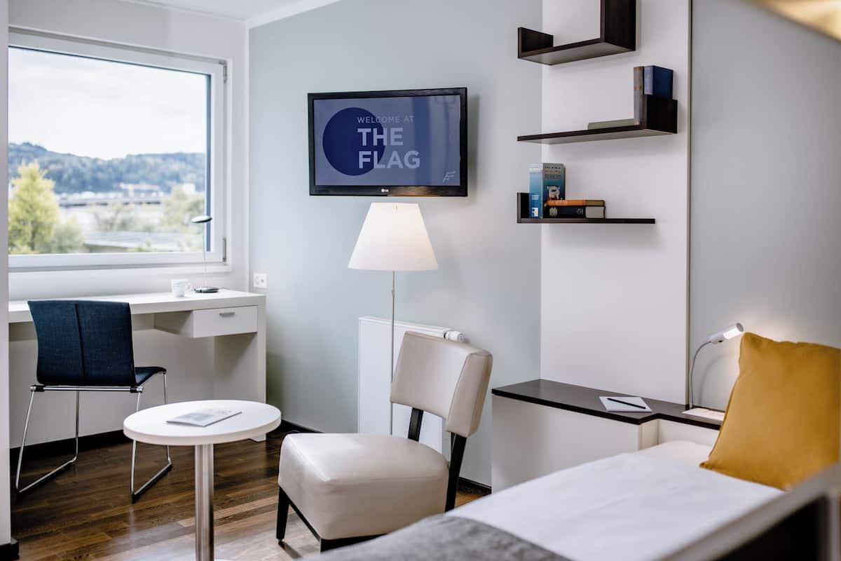 A hotel room with a desk, chair, bed, TV, lamp, and shelves with books.