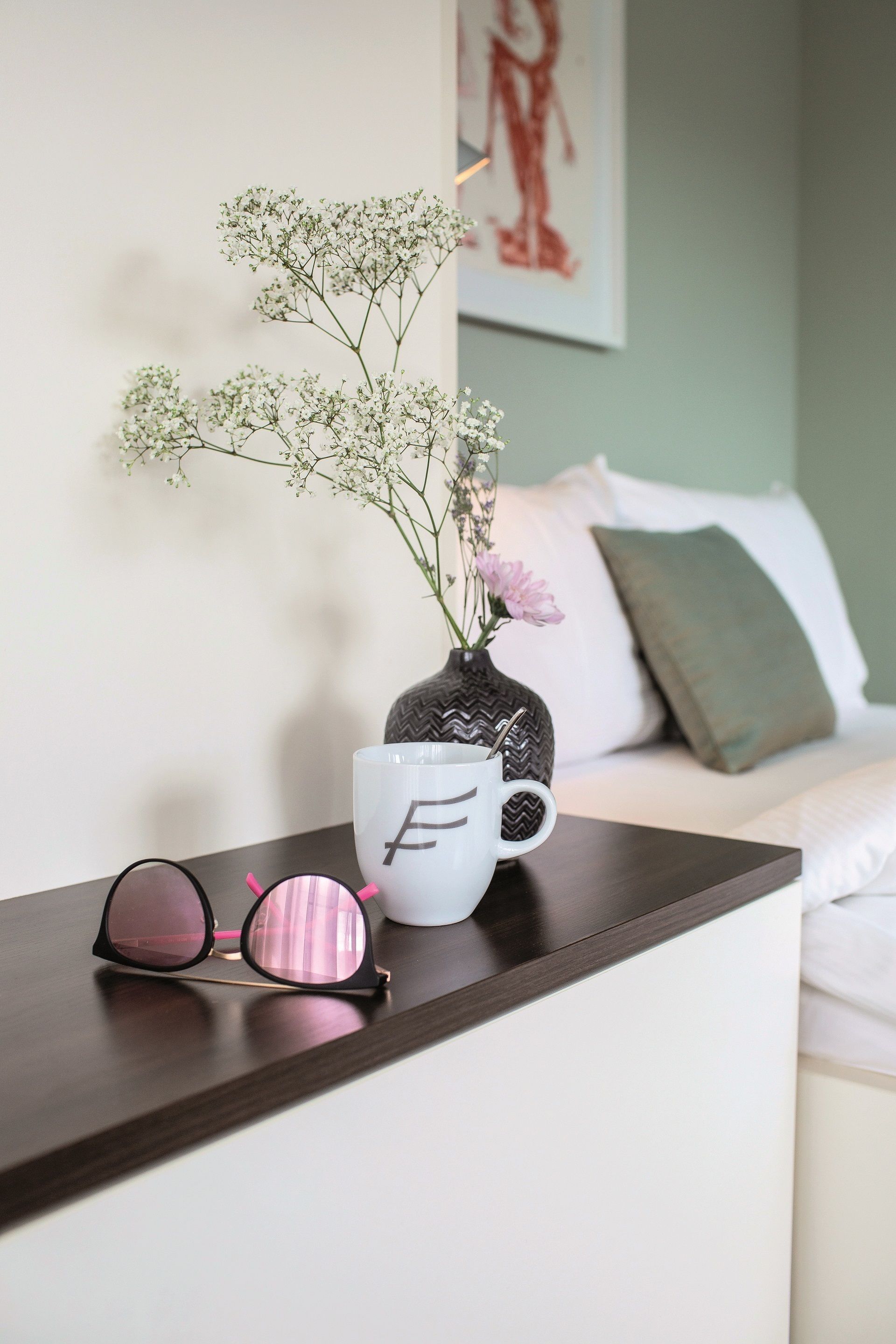 Una taza blanca con la letra 'F', unas gafas de sol rosas, y un florero con flores blancas y flores rosadas sobre una mesa de madera.