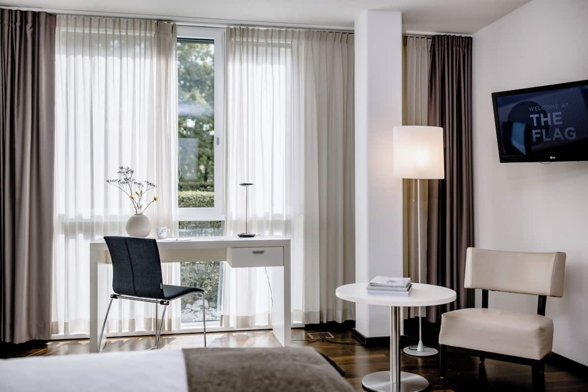 A hotel room with a bed, a desk with a chair, a small table with a lamp, and a TV on the wall.