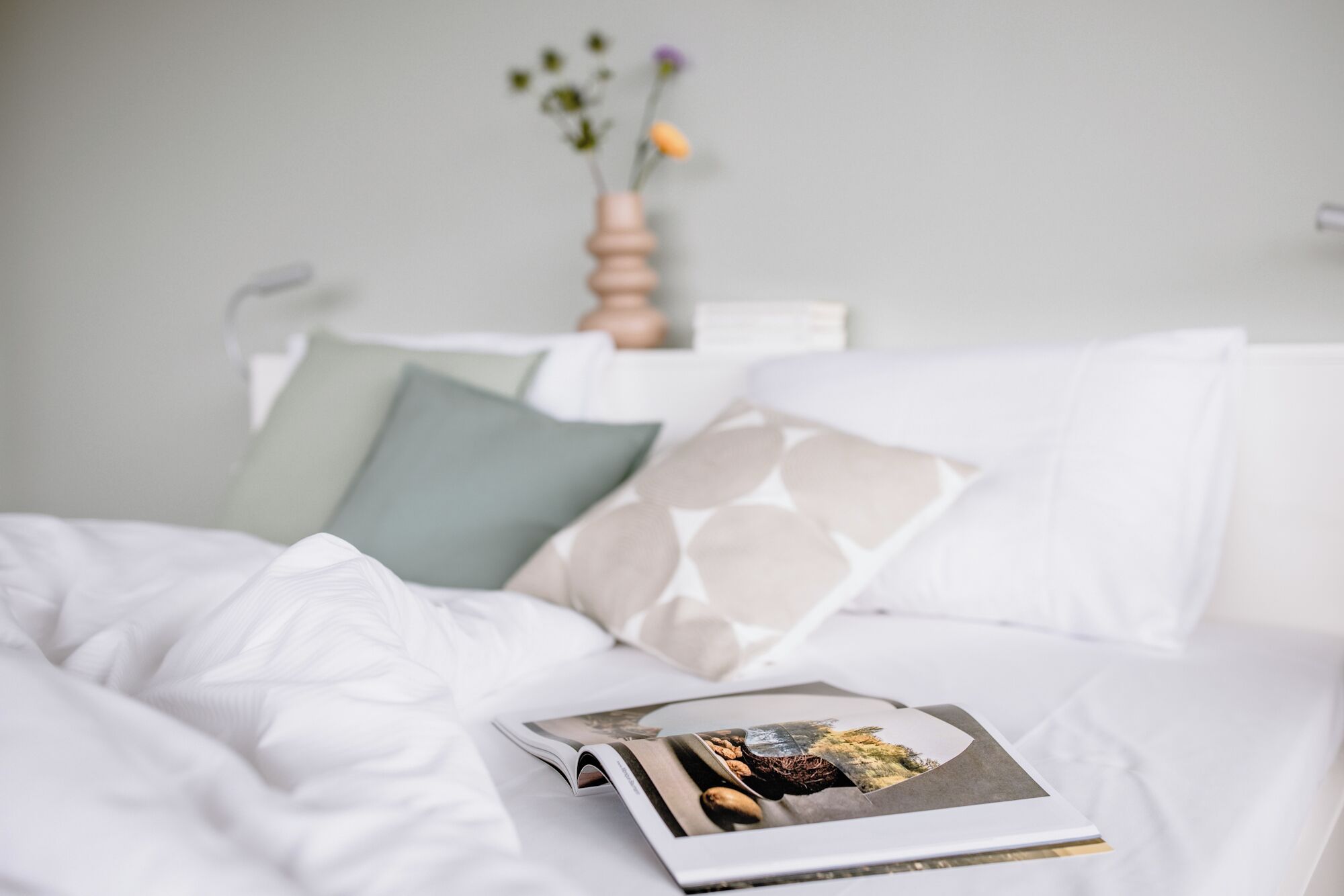 Una cama bien hecha con una revista encima, un florero con flores y una lámpara de pared.