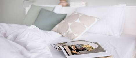 Una cama bien hecha con una revista encima, un florero con flores y una lámpara de pared.