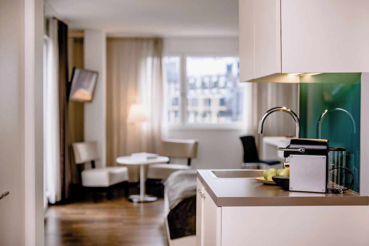 A modern hotel room with a kitchenette, a dining area, and a view of the city.