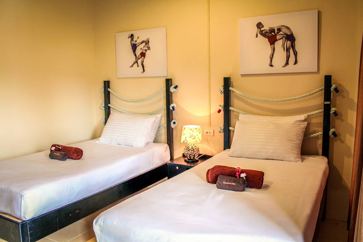 Two single beds with white linens, a nightstand with a lamp, and artwork depicting a boxing match on the walls.