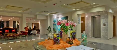 Un lobby espacioso con una mesa de vidrio, muebles rojos y dorados, y un cuadro de flores.