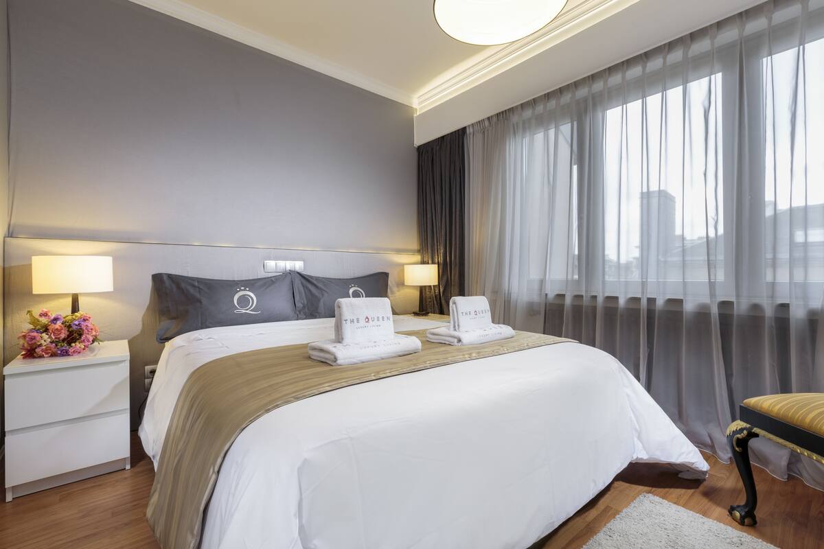A hotel room with a large bed, two bedside tables, a nightstand, a chair, and a view of the city through sheer curtains.
