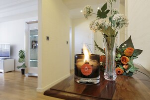 Una vela con llama encendida, un florero con flores y una mesa de madera en una habitación con piso de madera y pared blanca.